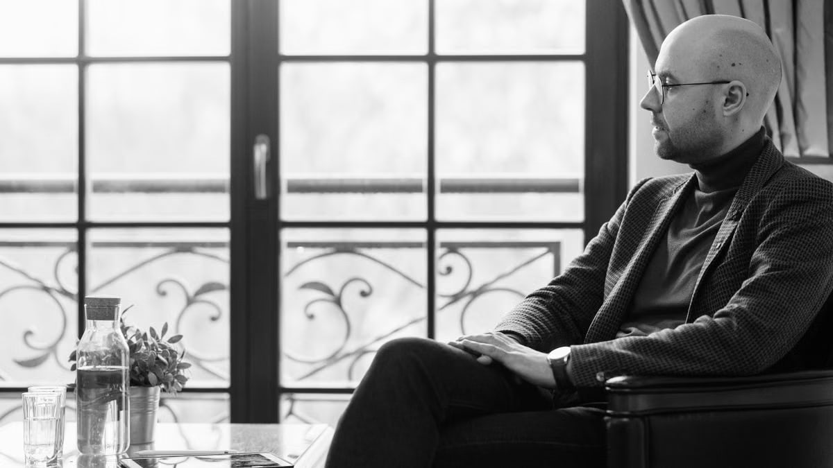 Black and white portrait of a bald man sitting thoughtfully near a window indoors.